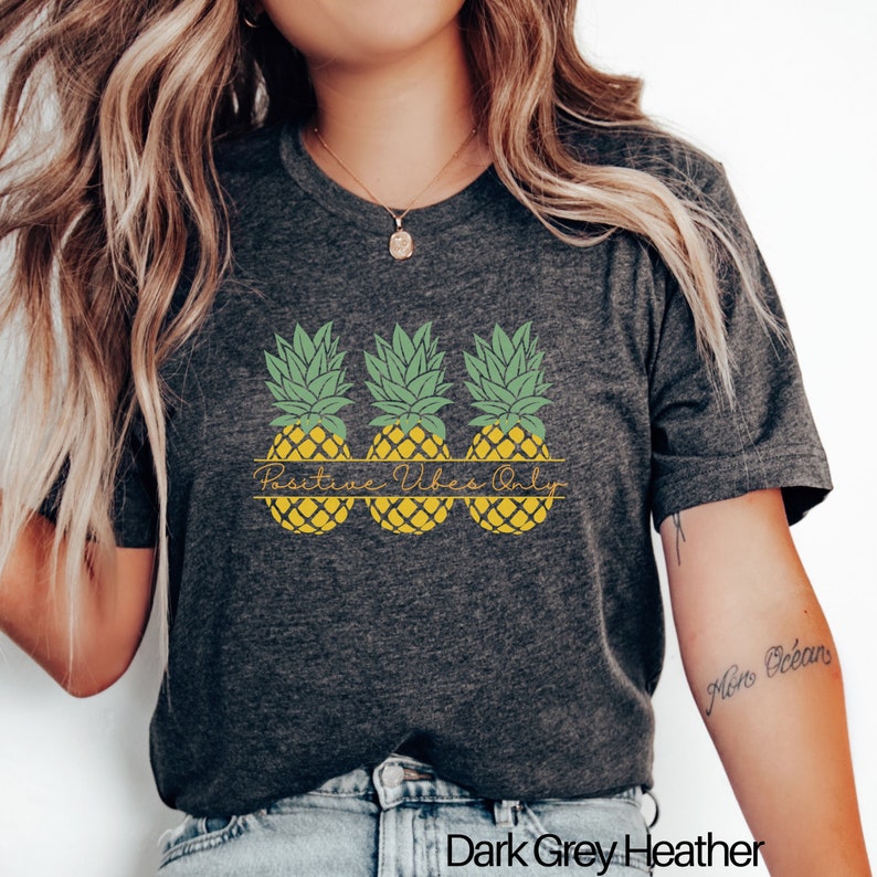 May include: A dark grey heather t-shirt with a graphic of three yellow pineapples with green tops. The text "Positive Vibes Only" is printed in a script font above the pineapples.
