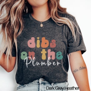 May include: A dark grey heather t-shirt with the text "Dibs on the Plumber" in colorful, rounded letters with hearts.
