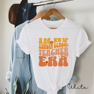 May include: White t-shirt with the text "In My Sunday School Teacher Era" in a retro font with orange and yellow colors and floral accents.