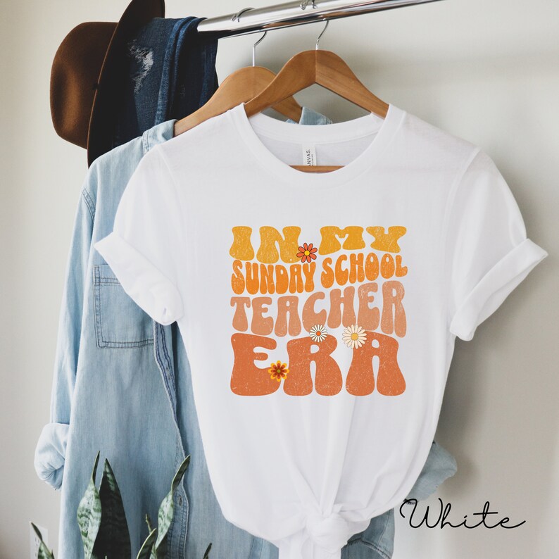 May include: White t-shirt with the text "In My Sunday School Teacher Era" in a retro font with orange and yellow colors and floral accents.
