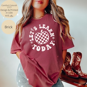May include: Brick-colored t-shirt with the text "LET'S LEARN TODAY" in a circular design around a checkered apple. The shirt is made of 100% cotton and is printed in the USA. The shirt is part of the Comfort Colors&reg; 1717 collection.