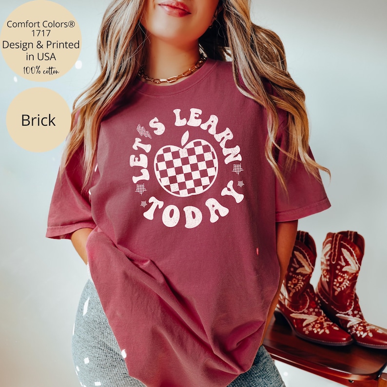May include: Brick-colored t-shirt with the text "LET'S LEARN TODAY" in a circular design around a checkered apple. The shirt is made of 100% cotton and is printed in the USA. The shirt is part of the Comfort Colors&reg; 1717 collection.