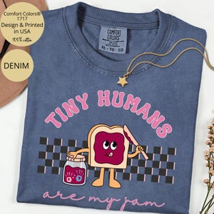 Pu&ograve; includere: T-shirt Comfort Colors blu denim con la scritta rosa "TINY HUMANS are my jam". La grafica presenta una fetta di pane tostato con marmellata, un barattolo di marmellata e un motivo a scacchiera.