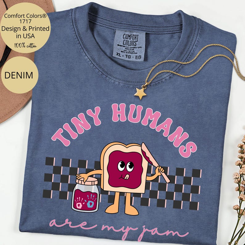 Pu&ograve; includere: T-shirt Comfort Colors blu denim con la scritta rosa "TINY HUMANS are my jam". La grafica presenta una fetta di pane tostato con marmellata, un barattolo di marmellata e un motivo a scacchiera.
