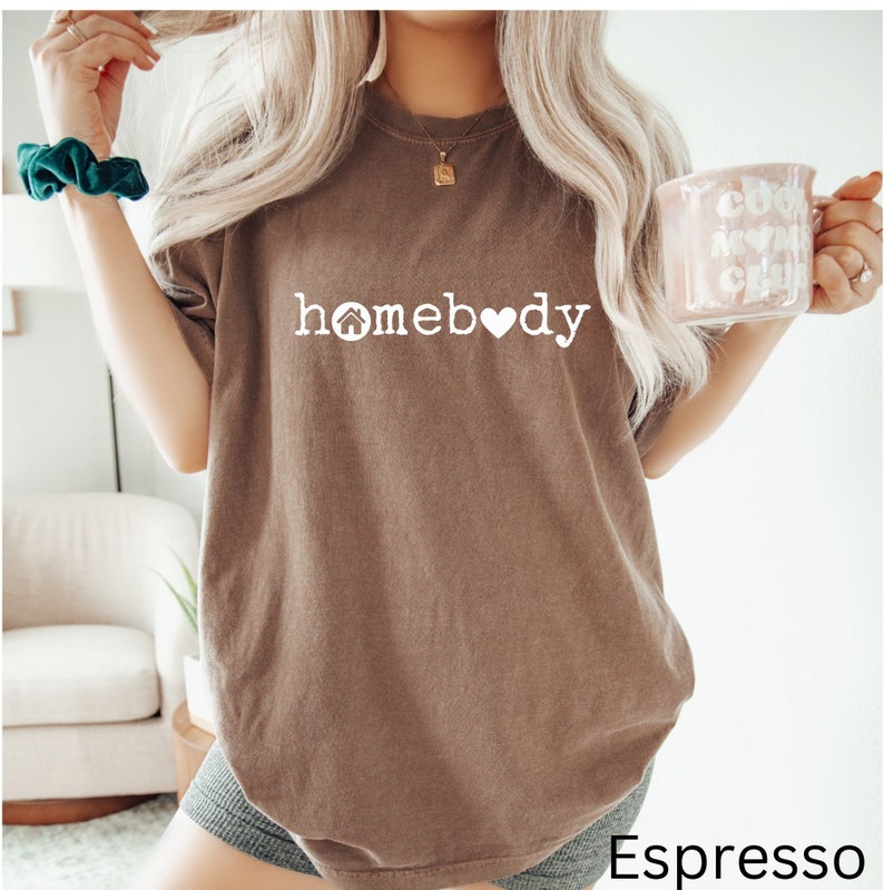 Pode incluir: Uma camiseta marrom oversized com o texto "homebody" em branco, com um &iacute;cone de casa e um &iacute;cone de cora&ccedil;&atilde;o.