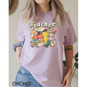 May include: A purple t-shirt with the word "Teacher" in a rainbow font. The shirt features a colorful graphic design with school-themed elements like a school bus, an apple, a lightning bolt, and a pencil.