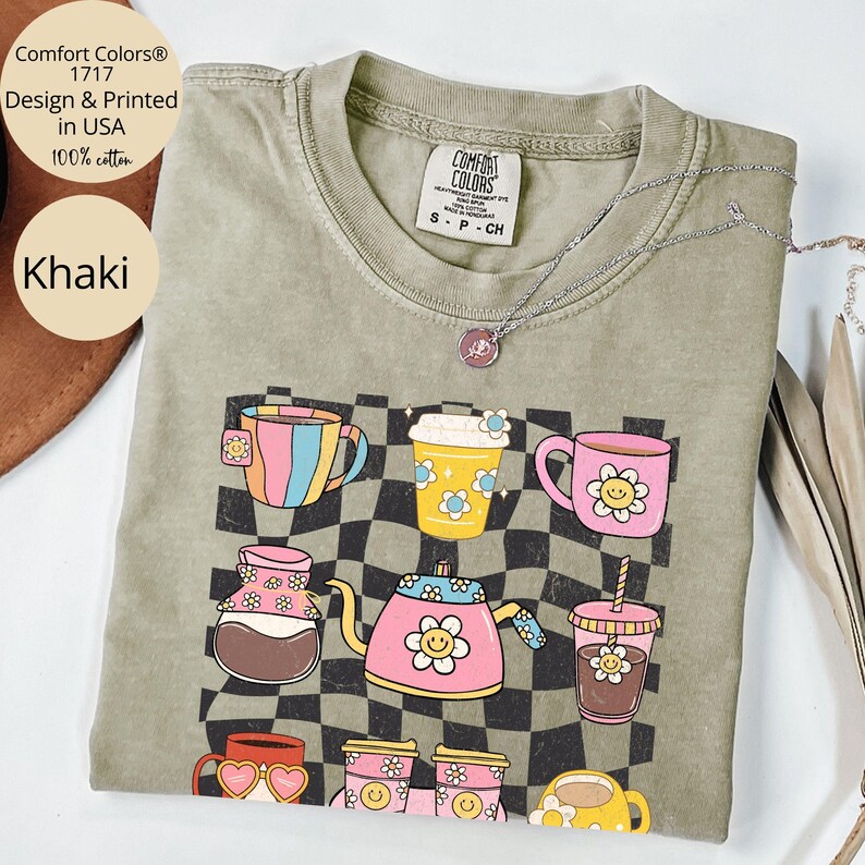 May include: Khaki t-shirt featuring a graphic print of assorted coffee mugs and cups against a checkered background. The text "Comfort Colors&reg; 1717 Design & Printed in USA 100% cotton" is printed in the top left corner of the shirt.
