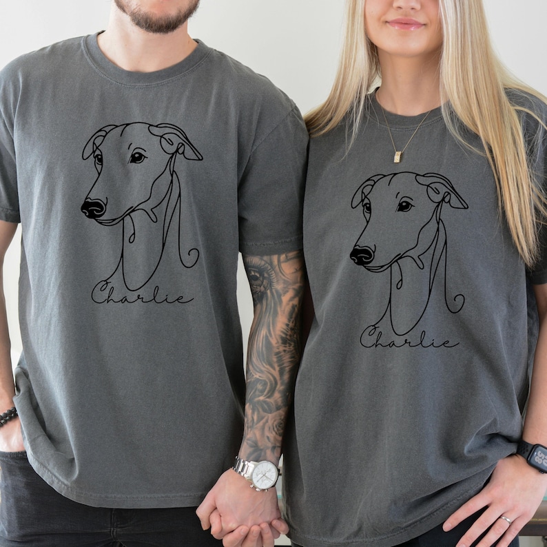May include: Two charcoal gray t-shirts featuring a minimalist line drawing of a dog's head. Below the drawing, the name "Charlie" is written in cursive. The shirts are a matching set, perfect for dog lovers.