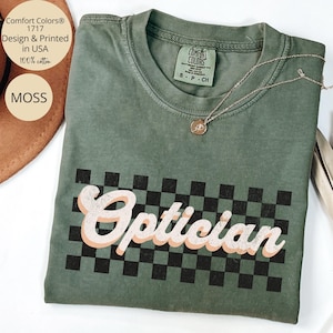 May include: A moss green crew neck t-shirt featuring a black and white checkered pattern graphic with the word Optician printed in a distressed white and peach script font across the centre. The shirt has a Comfort Colors brand label inside.