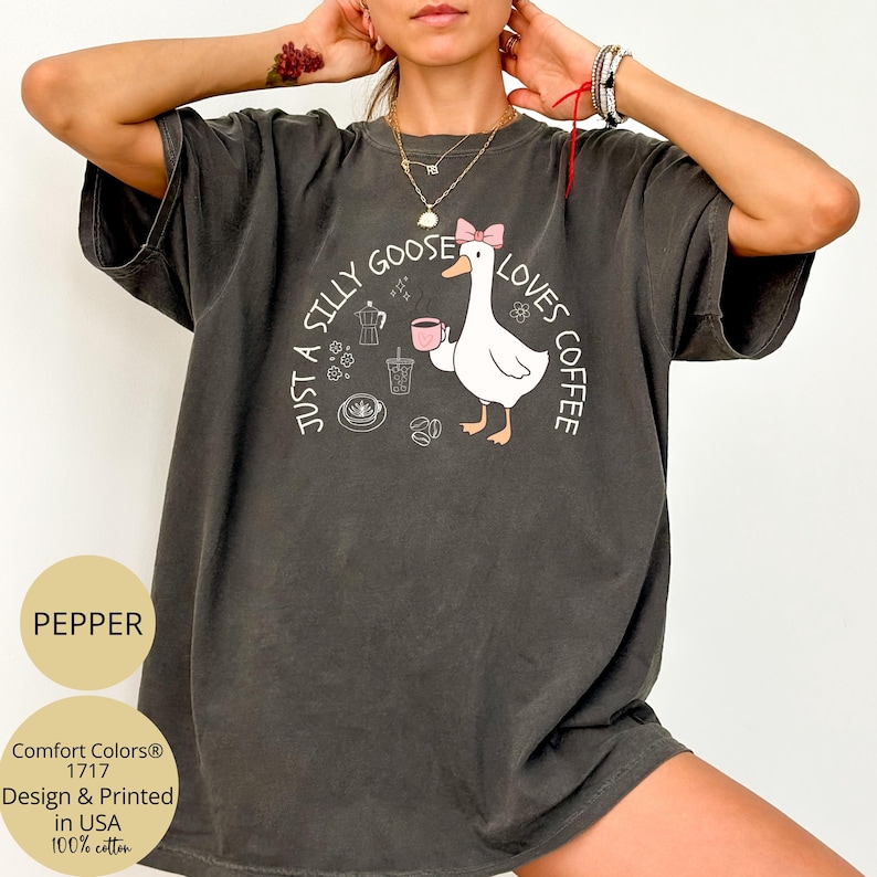 Puede incluir: Camiseta color pimienta con un gr&aacute;fico de un ganso con un lazo rosa, sosteniendo una taza de caf&eacute;, y el texto "Just a Silly Goose Loves Coffee." La camiseta es 100% algod&oacute;n.