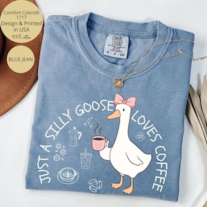 Puede incluir: Camiseta Comfort Colors azul vaquero con un dibujo de un ganso sosteniendo una taza de caf&eacute;, un lazo rosa y el texto "Just a Silly Goose Loves Coffee". La camiseta es 100% algod&oacute;n y est&aacute; dise&ntilde;ada e impresa en EE. UU.