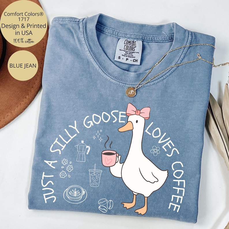 Puede incluir: Camiseta Comfort Colors azul vaquero con un dibujo de un ganso sosteniendo una taza de caf&eacute;, un lazo rosa y el texto "Just a Silly Goose Loves Coffee". La camiseta es 100% algod&oacute;n y est&aacute; dise&ntilde;ada e impresa en EE. UU.