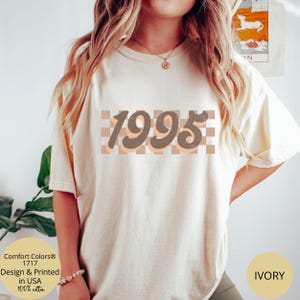May include: A white t-shirt with a brown and peach checkered design and the year 1995 in brown text. The text is in a retro font style. The t-shirt is available in ivory.