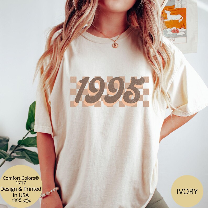 May include: A white t-shirt with a brown and peach checkered design and the year 1995 in brown text. The text is in a retro font style. The t-shirt is available in ivory.