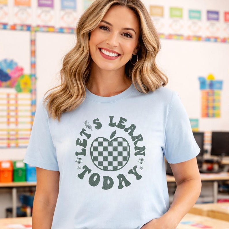 May include: Light blue t-shirt with the text "LET'S LEARN TODAY" in a circular design. The design includes a checkered apple in the center and stars. The shirt is short-sleeved and made of a soft material.
