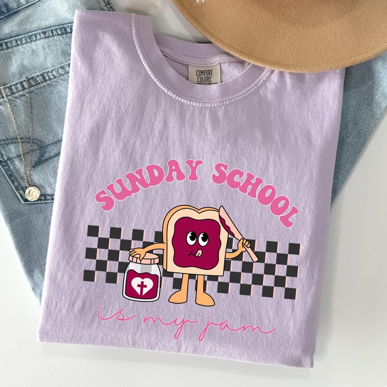 May include: A lavender t-shirt with a graphic of a cartoon slice of bread with a heart-shaped jar of jam. The text "SUNDAY SCHOOL is my jam" is printed on the shirt.