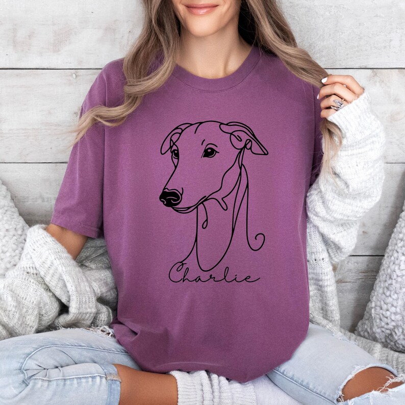 May include: A mauve-colored t-shirt featuring a minimalist line drawing of a dog's head. The dog's name, "Charlie," is written in a delicate script below the drawing. The shirt has a relaxed fit and is made of soft material.