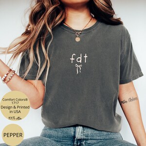Peut inclure: Un t-shirt gris fonc&eacute; avec les lettres "fdt" et un motif de n&oelig;ud blanc. Le t-shirt est en 100% coton et est imprim&eacute; aux &Eacute;tats-Unis. Le t-shirt est un mod&egrave;le Comfort Colors 1717 de couleur Pepper.