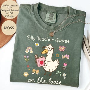 Pode incluir: Uma camiseta verde com o texto "Silly Teacher Goose on the loose" em branco. Um desenho animado de um ganso de &oacute;culos e segurando um livro com uma ma&ccedil;&atilde; na capa est&aacute; no centro da camiseta. O ganso est&aacute; rodeado de ilustra&ccedil;&otilde;es coloridas de flores, arco-&iacute;ris, estrelas e la&ccedil;os.