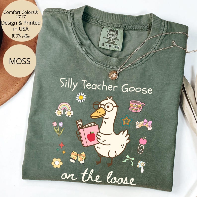 Pode incluir: Uma camiseta verde com o texto "Silly Teacher Goose on the loose" em branco. Um desenho animado de um ganso de &oacute;culos e segurando um livro com uma ma&ccedil;&atilde; na capa est&aacute; no centro da camiseta. O ganso est&aacute; rodeado de ilustra&ccedil;&otilde;es coloridas de flores, arco-&iacute;ris, estrelas e la&ccedil;os.