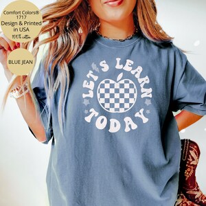 May include: Blue jean Comfort Colors t-shirt with the text "LET'S LEARN TODAY" in white. The design includes a checkered apple graphic. The shirt is made of 100% cotton and is designed and printed in the USA.