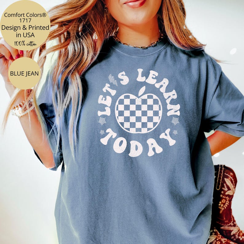 May include: Blue jean Comfort Colors t-shirt with the text "LET'S LEARN TODAY" in white. The design includes a checkered apple graphic. The shirt is made of 100% cotton and is designed and printed in the USA.