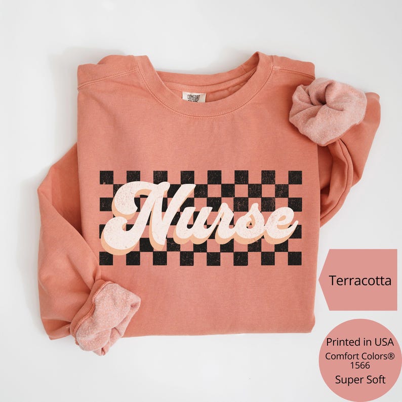 K&ouml;nnte beinhalten: Terracottafarbenes Sweatshirt mit dem Wort "Nurse" in Retro-Schrift &uuml;ber einem schwarz-wei&szlig;en Schachbrettmuster. Das Sweatshirt hat lange &Auml;rmel und ein passendes Scrunchie. Der Text "Printed in USA Comfort Colors&reg; 1566 Super Soft" ist ebenfalls sichtbar.