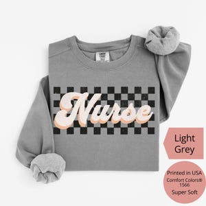 K&ouml;nnte beinhalten: Hellgraues Sweatshirt mit dem Wort "Nurse" in einem Retro-Schriftzug mit Pfirsich-Umriss, vor einem schwarz-wei&szlig; karierten Hintergrund. Das Sweatshirt ist superweich und in den USA bedruckt.