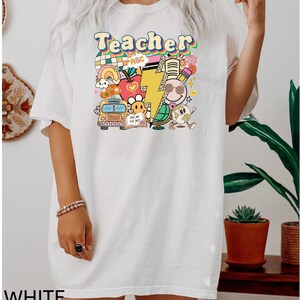 May include: White oversized t-shirt with a graphic print of a school bus, apple, lightning bolt, globe, and the word "Teacher" in rainbow colors.