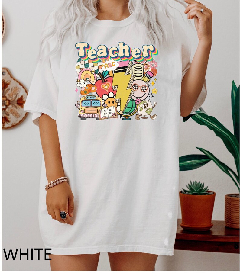 May include: White oversized t-shirt with a graphic print of a school bus, apple, lightning bolt, globe, and the word "Teacher" in rainbow colors.