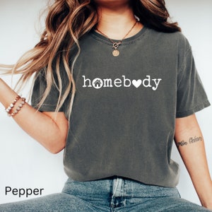 Pode incluir: Uma camiseta cinza escura com o texto "homebody" em branco com um pequeno &iacute;cone de casa e um &iacute;cone de cora&ccedil;&atilde;o.
