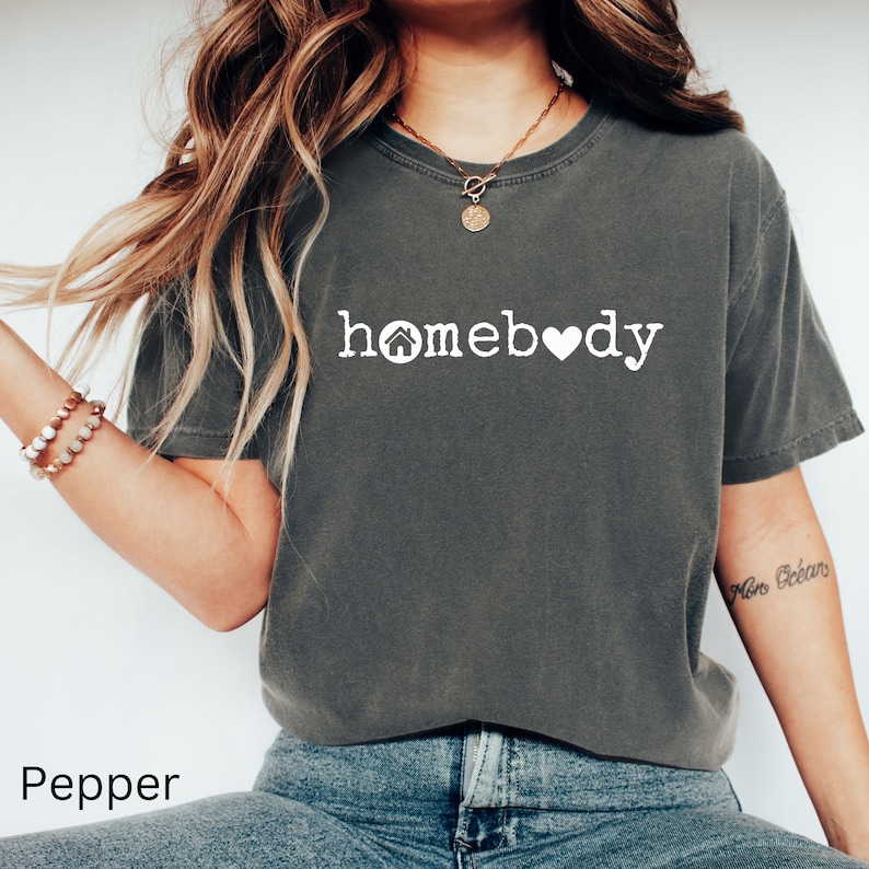 Pode incluir: Uma camiseta cinza escura com o texto "homebody" em branco com um pequeno &iacute;cone de casa e um &iacute;cone de cora&ccedil;&atilde;o.