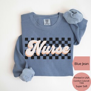 K&ouml;nnte beinhalten: Ein blaues Jeans-Sweatshirt mit dem Wort "Nurse" in einem Retro-Schriftzug &uuml;ber einem schwarz-wei&szlig;en Schachbrettmuster. Das Sweatshirt ist ein Comfort Colors 1566 Super Soft. Der Text "Printed in USA" ist ebenfalls sichtbar.
