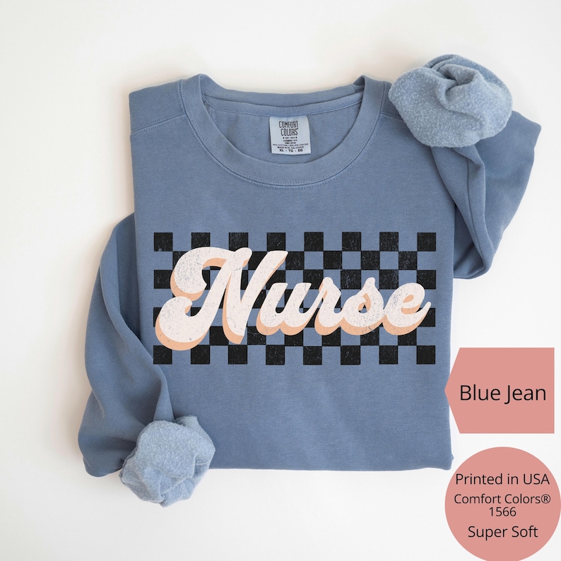 K&ouml;nnte beinhalten: Ein blaues Jeans-Sweatshirt mit dem Wort "Nurse" in einem Retro-Schriftzug &uuml;ber einem schwarz-wei&szlig;en Schachbrettmuster. Das Sweatshirt ist ein Comfort Colors 1566 Super Soft. Der Text "Printed in USA" ist ebenfalls sichtbar.