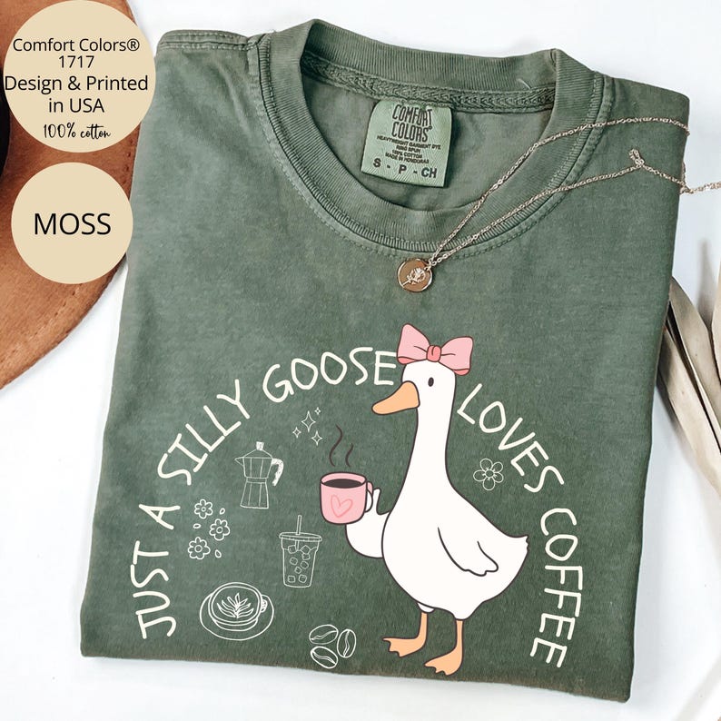 Puede incluir: Camiseta Comfort Colors verde musgo con un gr&aacute;fico de un ganso sosteniendo una taza de caf&eacute;, el texto dice "Just a Silly Goose Loves Coffee". La camiseta es 100% algod&oacute;n e impresa en EE. UU. Tambi&eacute;n se ve un collar plateado con un colgante.