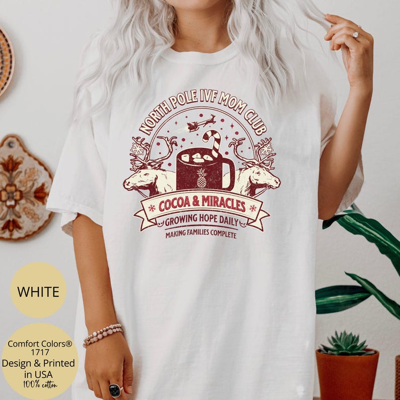 Puede incluir: Camiseta blanca con un dise&ntilde;o gr&aacute;fico rojo que dice "North Pole IVF Mom Club" con una ilustraci&oacute;n de una taza de cacao y un reno. El texto debajo del gr&aacute;fico dice "Cocoa & Miracles", "Growing Hope Daily" y "Making Families Complete".