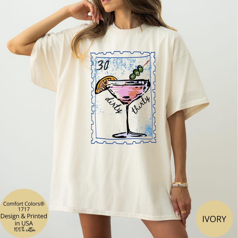 May include: Ivory-colored t-shirt featuring a cocktail design within a blue postage stamp frame. The design includes a pink drink, an orange slice, and olives. Text on the shirt reads "dirty thirty" and "30". Made of 100% cotton.