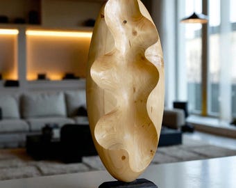 May include: A light-colored wooden sculpture with a flowing, organic design. The sculpture is oval-shaped and features a carved, wavy pattern down the center. It stands on a small, rectangular black base. The artwork is displayed indoors, suggesting a focus on home decor.