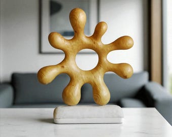 Könnte beinhalten: Helle Holzskulptur mit organischer Form, einem zentralen Loch und abgerundeten Ausläufern. Sie steht auf einem weißen, rechteckigen Sockel. Ein modernes Dekorationselement für ein zeitgemäßes Interieur.