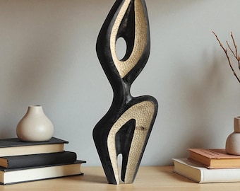 May include: A tall, abstract wooden sculpture with a black exterior and a textured, light beige interior. The sculpture has a flowing, organic shape with open spaces and a smooth finish. It stands on a wooden surface, suggesting a decorative home accent.
