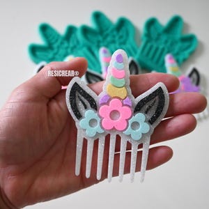 May include: A glittery white unicorn hair comb with a rainbow horn, black ears, and pink and blue flower accents. The comb is held in a hand, with other similar combs in the background. The brand name "RESICREAR" is visible.