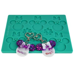 May include: Two Minnie Mouse-themed keychains with glitter accents and the names "Sophia" and "Stacy" on white circles. They are displayed on a teal silicone mold with Mickey Mouse and bow shapes. The keychains have silver heart-shaped clasps.