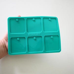 May include: A teal silicone mold with six square cavities, each with a small circular indentation. The mold has the text "RESICREAR®" printed on it. The mold is held against a white background.