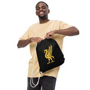 May include: A black drawstring bag with a gold Liverpool Football Club crest. The bag is being held by a person wearing a beige t-shirt.