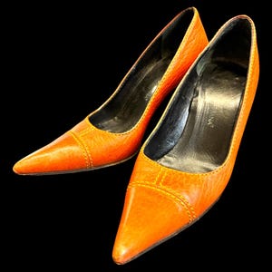 May include: A pair of orange leather pumps with pointed toes. The shoes have a low heel and a stitched design around the toe box. The interior is black.