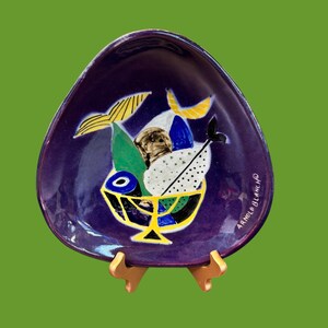 May include: A deep purple, teardrop-shaped ceramic dish with a painted still life of fruit in a yellow bowl. The fruit includes bananas, a green leaf, and a white dotted fruit. The dish is signed "ARNOLD BLANCH".