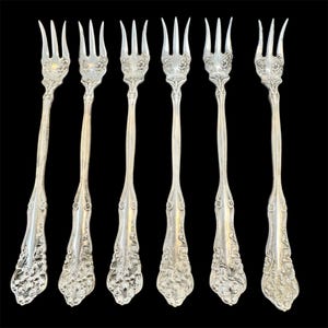 May include: Six silver-colored forks with ornate floral designs on the handles and tines. The forks are arranged in a row against a black background, showcasing their intricate details and elegant style. The forks are ideal for serving appetizers.