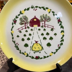May include: A vintage decorative plate with a yellow rim and a central farm scene. The scene features a red barn, green trees, a hay bale, and a path, all encircled by a green and fruit-laden wreath. The plate is displayed on a black stand.