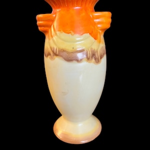 May include: A vintage ceramic vase with a cream-colored body and a flared orange top. The vase features two side handles and a decorative band of yellow and brown near the top. The base is a rounded shape. The vase is approximately 10 inches tall.