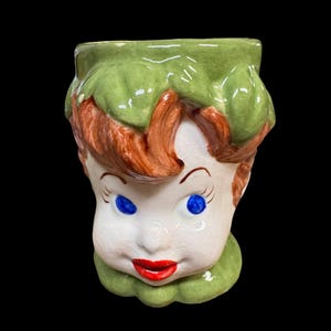 May include: A ceramic head-shaped planter or mug featuring a glossy finish with a crackle texture. The figure has bright blue eyes, red lips, and brown hair styled under a green leafy cap, with a matching green leafy base at the bottom.